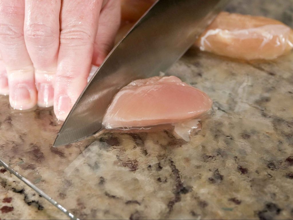 Nahaufnahme einer Hand, die ein rohes Hähnchenfilet auf einem Glasschneidebrett mit einem Messer schneidet.