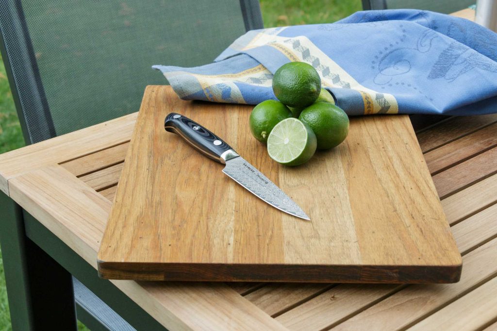 Holzschneidebrett auf Gartentisch mit Messer, mehreren Limetten und blau-weiß gemustertem Geschirrtuch.