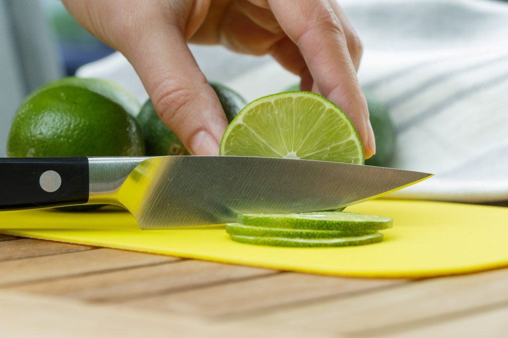 Hand schneidet Limette mit Messer in dünne Scheiben auf gelbem Plastikschneidebrett.