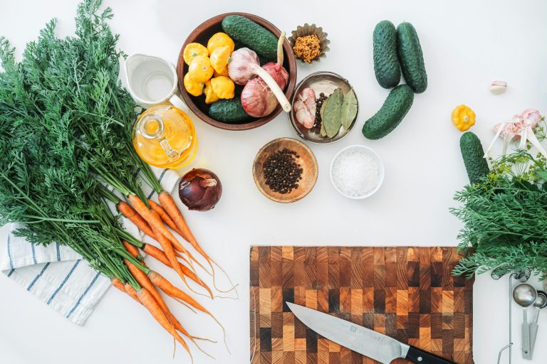 Karotten mit Grün, Gurken, Knoblauch, Zwiebel, gelbe Paprika, Gewürze in Schalen, Öl und ein Messer auf einem Schneidebrett auf weißem Untergrund